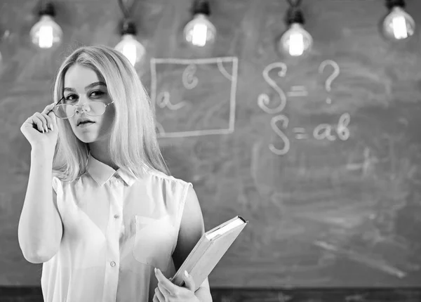 El profesor se ve confiado en las gafas, de pie en el aula, pizarra en el fondo. La mujer con libro comienza la lección, mira al público mientras se quita las gafas. Atractivo concepto de profesor — Foto de Stock