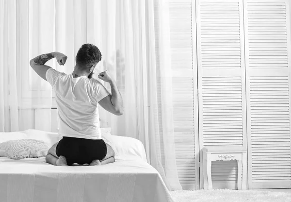 Tipo estirando los brazos, lleno de energía en la mañana, vista trasera. Concepto matutino perfecto. El hombre de camisa se sienta en la cama, cortinas blancas en el fondo. Macho en ropa interior estirándose, relajándose después de la siesta, descansar —  Fotos de Stock