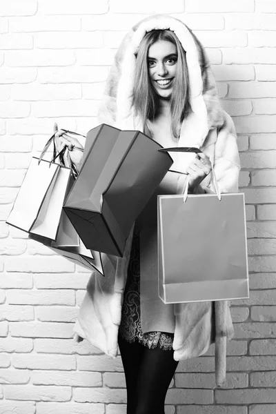 Jeune femme sexy jolie à la mode ou fille avec de longs beaux cheveux blonds en robe rose manteau de fourrure blanche avec capuche et maquillage de mode tenant paquet coloré sur fond de studio de mur de brique — Photo