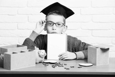 Genç Sevimli öğrenci çocuk gri kazak ve sınıf mezuniyet şapkalı beyaz tuğla duvar arka planda, kopya spac boşaltmak el ahşap numaraları pembe domuz domuz banka ile masa başında oturan gözlük