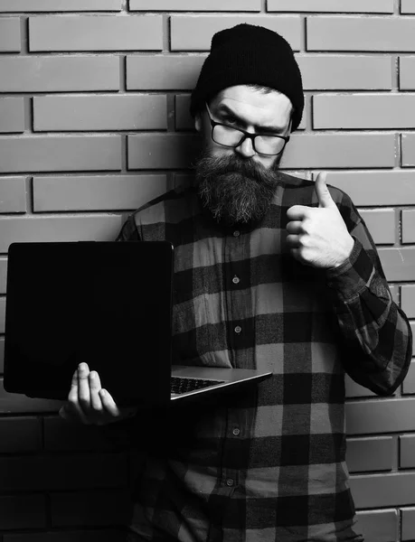 Sakallı adam, uzun sakal. Acımasız beyaz ciddi tıraşsız hipster dizüstü şapka ve gözlük kahverengi tuğla duvar studio adam kırmızı siyah damalı gömlek holding — Stok fotoğraf