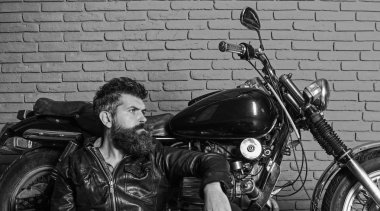 Sakallı, motorcu motorlu bisiklet garaj, tuğla duvar arka plan yakınındaki deri ceketli adam. Yenilikçi, vahşi motorcu deri ceketli dalgın yüzünde oturuyor, motosiklet üzerinde eğilir. Bisikletçileri yaşam tarzı kavramı