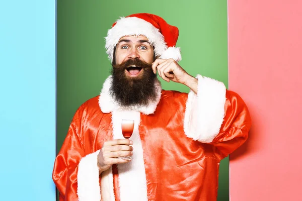 Knappe bebaarde kerstman met lange baard op een glimlachend gezicht met een glas alcoholische shot in rode kerst- of kerstjas en nieuwjaarshoed op een kleurrijke studioachtergrond — Stockfoto