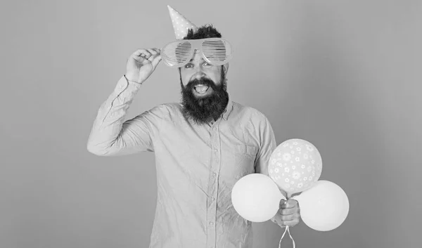 Homem barbudo alegre com grande sorriso usando óculos enormes e boné de aniversário. Hipster feliz com barba aparada e bigode em camisa rosa posando com balões amarelos e verdes em fundo azul — Fotografia de Stock