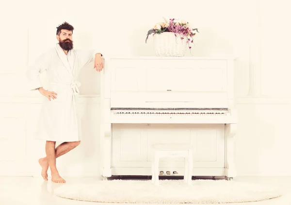 Homem com barba em roupão goza de manhã enquanto está perto do piano. Conceito de músico talentoso. Homem sério fica e se inclina no piano instrumento musical em interior branco no fundo — Fotografia de Stock