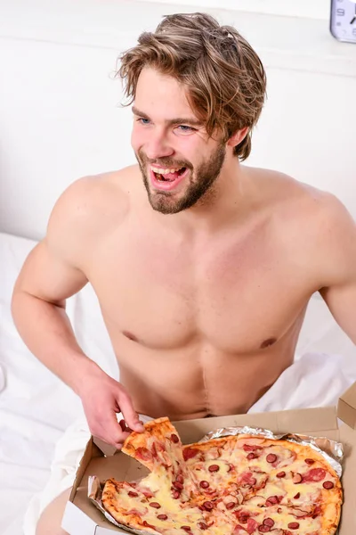 Shirtless handsome young man with pizza on bed. Guy holds pizza box sit bed in bedroom or hotel room. Portrait of lazy muscular man eating pizza while laying on a bed at home. Pizza on bed. — Stock Photo, Image