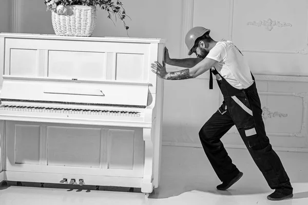 Heavy loads concept. Loader moves piano instrument. Courier delivers furniture, move out, relocation. Man with beard worker in helmet and overalls pushes, put efforts to move piano, white background — Stock Photo, Image