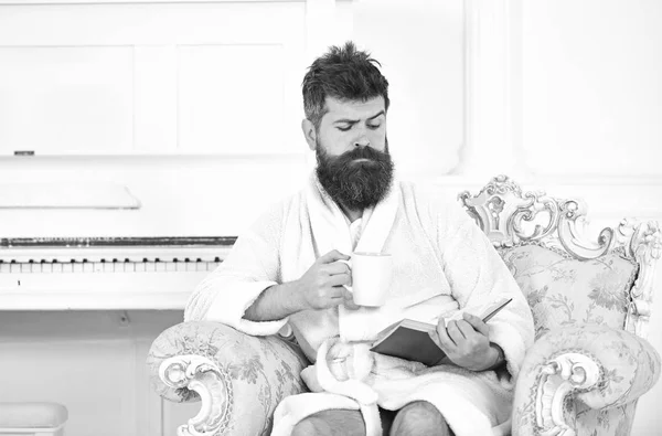 Человек с бородой и усами наслаждается утром, сидя на роскошном кресле. Мужчина сонный в халате, пьет кофе, читает книгу в роскошном отеле утром, белый фон. Элитный досуг — стоковое фото