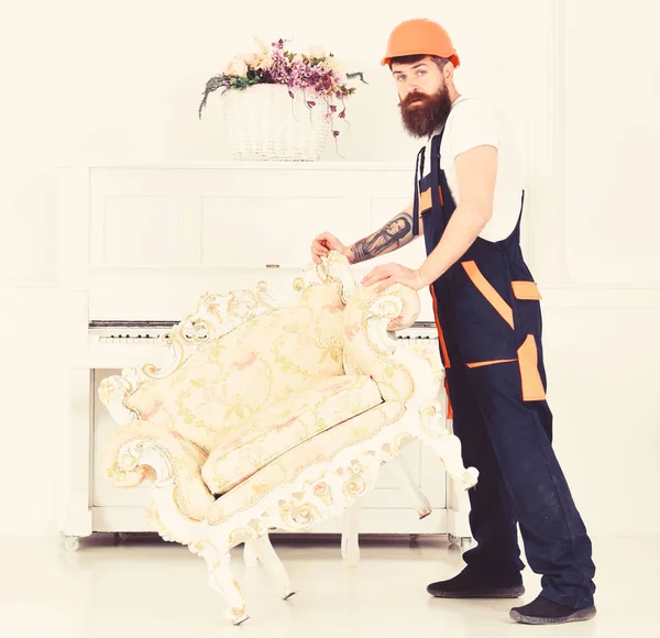 Relocating concept. Courier delivers furniture in case of move out, relocation. Man with beard, worker in overalls and helmet lifts up armchair, white background. Loader moves armchair for move out