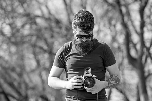 Skäggig man arbetar med vintage kamera. Mannen med skägg och mustasch på koncentrerade ansikte, grenar på bakgrund, oskärpa. Fotograf koncept. Man med långt skägg upptagen med fotografering — Stockfoto