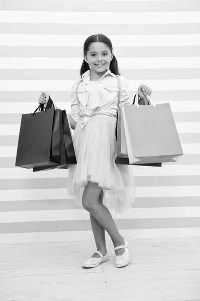 Zakupy jest jej ulubionym zajęciem. Dziecko dziewczynka prowadzi bacground torby na zakupy w paski. W każdym wieku kobiety uwielbiają zakupy. Dziecko dziewczynka zakupy sezon sprzedaż. Dziewczyna uśmiecha się zadowolony twarz posiada torby — Zdjęcie stockowe
