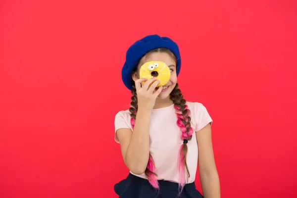 Um miúdo fã de donuts assados. Delicioso donut doce. Menina em chapéu de boina segurar donut fundo vermelho. Miúdo menina brincalhão comer donut. Conceito de saúde e nutrição. Bela vida. Loja de doces e conceito de padaria — Fotografia de Stock