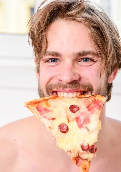 Serviço de entrega de alimentos. Bonito homem segura um pedaço de pizza em suas mãos e está prestes a comê-lo. Guy detém caixa de pizza sentar cama no quarto ou quarto de hotel. Refeição . — Fotografia de Stock