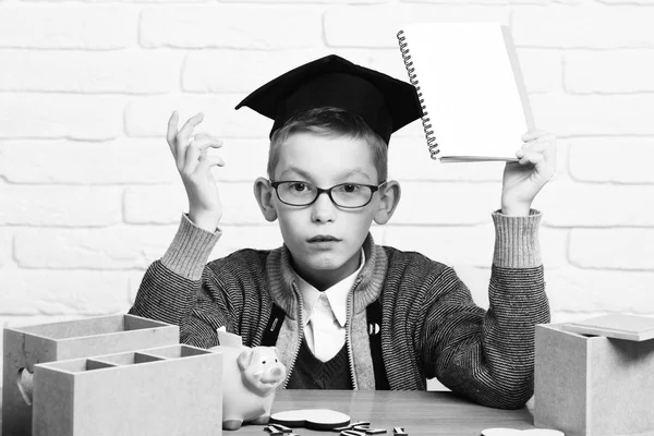 Unga söta elev pojke i grå tröja och glasögon som sitter vid skrivbord med häfte i hand trä siffror rosa grisen gris bank i examen keps i klassrummet på vitt tegel vägg bakgrund — Stockfoto