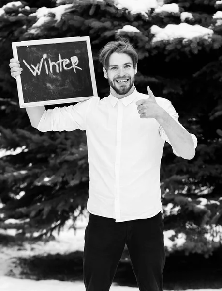 Красивый бородатый мужчина или сексуальный улыбающийся счастливый парень в белой рубашке держит доску или доску с зимним текстом, показывает большой палец вверх на зеленом елке на естественном фоне, копия spac — стоковое фото