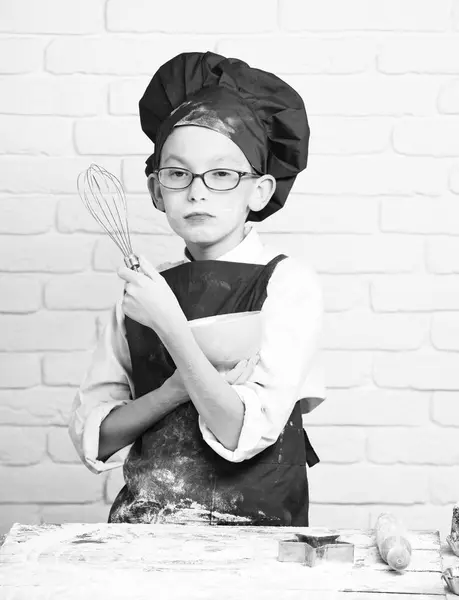 Young boy cute cook chef in red uniform and hat on stained face flour with glasses standing near table with rolling pin and holding kitchen whisk and turquoise bowl on white brick wall background — Stock Photo, Image