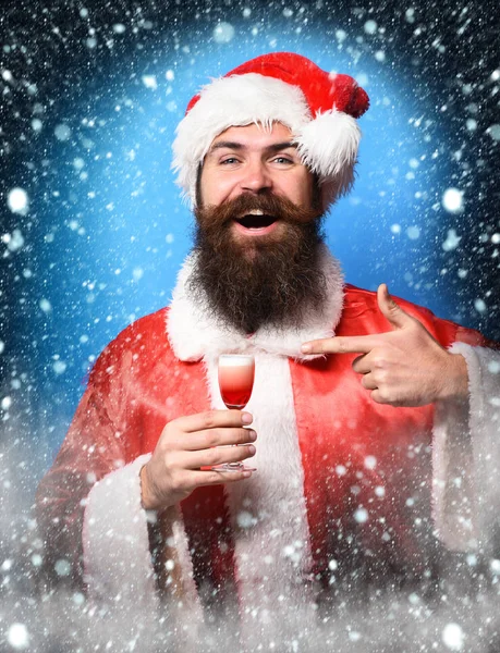 handsome bearded santa claus man with long beard on smiling face holding glass of alcoholic shot in red christmas or xmas sweater and new year hat on blue studio background