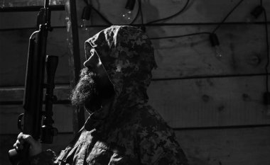 Sakallı adam başlıklı kamuflaj elbiseler, ahşap iç arka plan giyer. Avcı kavramı. Hunter, hipster silahla tüfek avcılık için hazırlayın. Maço gamekeepers evinde, kopya spac sıkı yüzünde