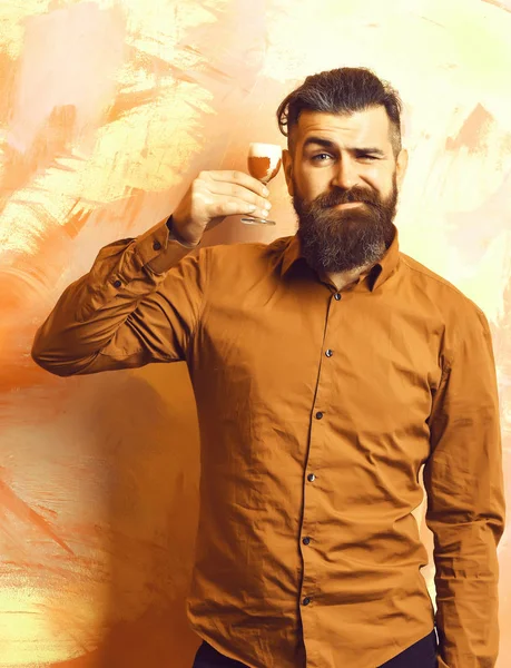 Bearded man, long beard. Brutal caucasian happy hipster with moustache in brown shirt holding alcoholic red shot on colorful texture backgroun