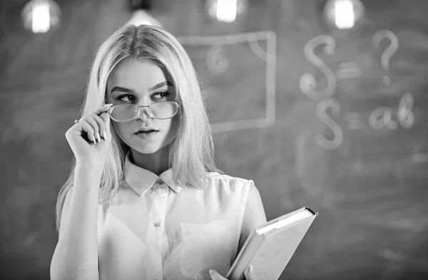 Concept de conférencier attrayant. Femme avec livre commence la leçon, regarde le public tout en enlevant les lunettes. Professeur semble confiant dans les lunettes, debout dans la salle de classe, tableau en arrière-plan — Photo