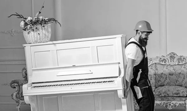 Loader moves piano instrument. Man with beard, worker in overalls and helmet lifts up piano, white background. Courier delivers furniture in case of move out, relocation. Delivery service concept
