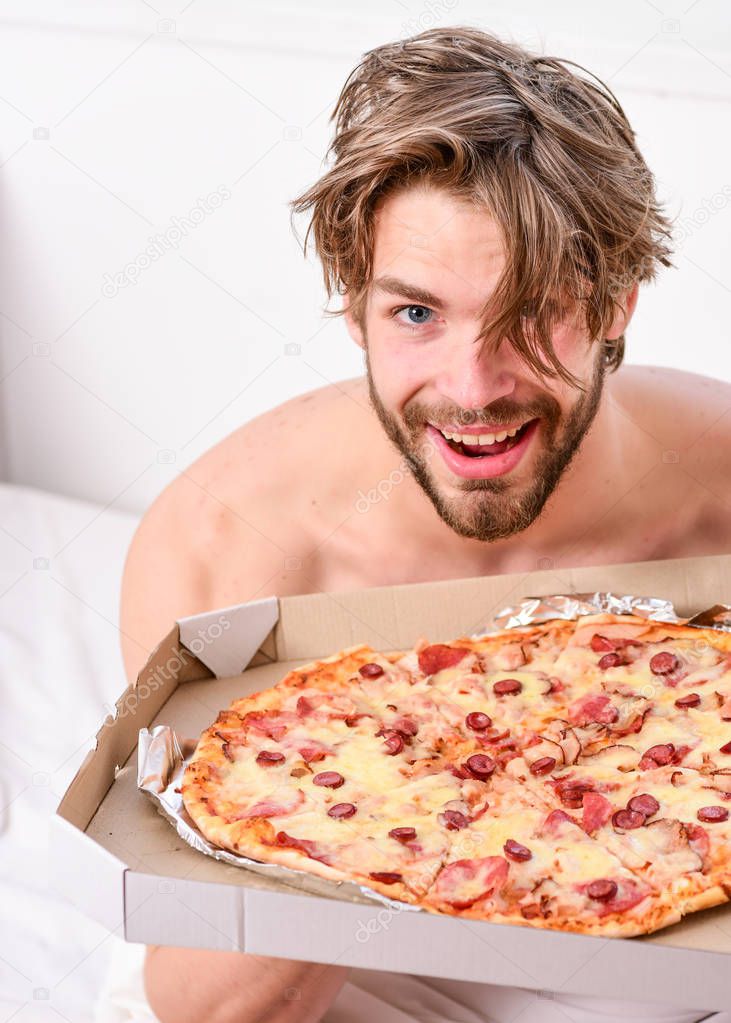 Man likes pizza for breakfast. Sexy man eat pizza lying on bed. Portrait of lazy muscular man eating pizza while laying on a bed at home. Home pizza.