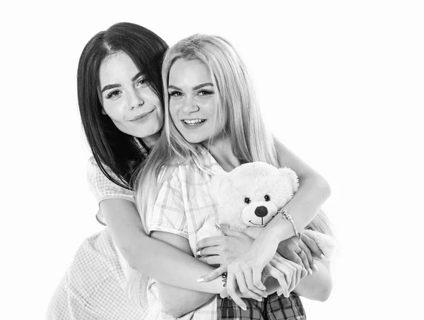 Rubia y morena en caras sonrientes en ropa para dormir se ve linda y amigable. Chicas abrazándose, aisladas sobre fondo blanco. Hermanas o mejores amigas en pijama con peluche oso de juguete. Lindo concepto — Foto de Stock