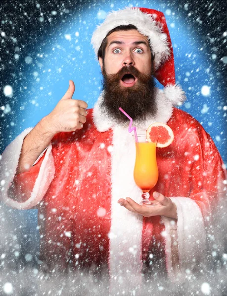 Schöner bärtiger Weihnachtsmann mit langem Bart im überraschten Gesicht, der ein Glas alkoholfreien Cocktails in Weihnachts- oder Weihnachtspullover und Neujahrsmütze auf blauem Studiohintergrund hält — Stockfoto