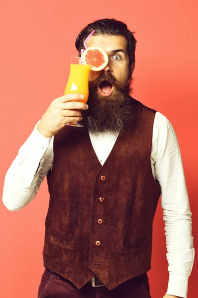 Handsome bearded man with long beard and mustache has stylish hair on surprised face holding glass of alcoholic cocktail in vintage suede leather waistcoat on red studio background — Stock Photo, Image