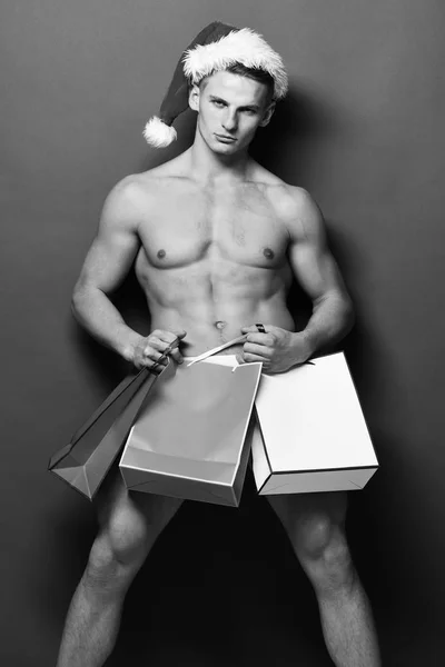 Mladý pohledný macho santa muž se sexy svalnatý sportovní silné tělo má holé torzo a silné břicho v Vánoce nebo Vánoce klobouk drží barevné přítomných sáčků na zelené studio pozadí — Stock fotografie