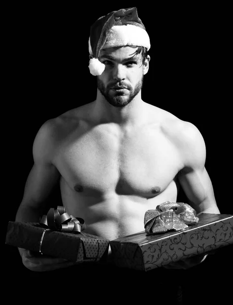 handsome bearded man or sexy christmas guy with muscular body, chest and torso in red santa claus hat holds present or gift boxes for new year or xmas holiday celebration on black backgroun