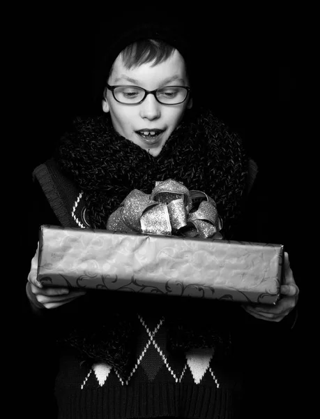 Pequeño niño sonriente o lindo niño nerd en gafas, sombrero y bufanda de punto de moda sobre fondo negro sostiene caja de regalo rojo con oro bo —  Fotos de Stock