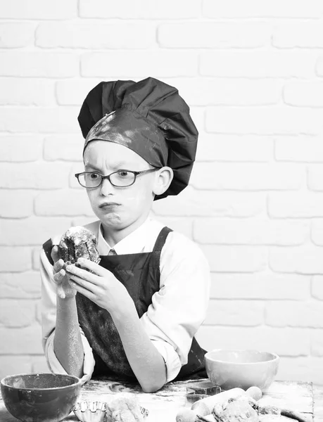 Young boy cute cook chef in red uniform and hat on stained face with glasses sitting on table with colorful bowls, tasty cookies, rolling pin and holding chocolate cake on white brick wall backgroun — Stock Photo, Image