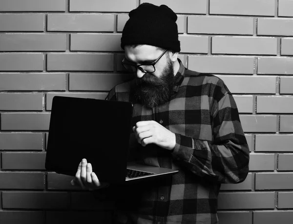 Bearded man, long beard. Brutal caucasian serious unshaven hipster holding laptop in red black checkered shirt with hat and glasses on brown brick wall studio backgroun