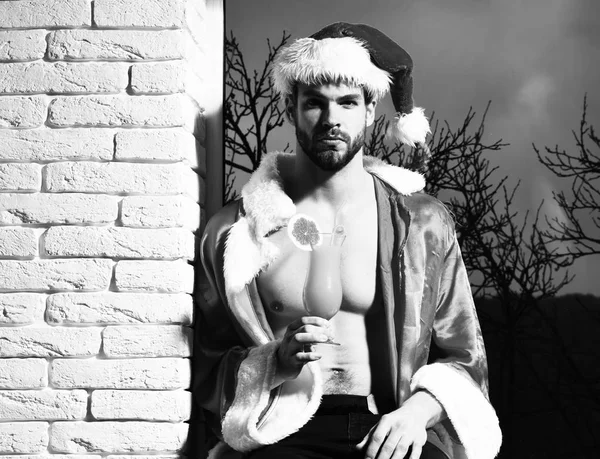 Mladý pohledný vousatý sexy vánoční muž se stylovým vousem v červené Santa Claus klobouk a kabát s svalnatý holý trup drží sklo alkoholického pomerančového nápoje na černém a cihlové zdi pozadí — Stock fotografie