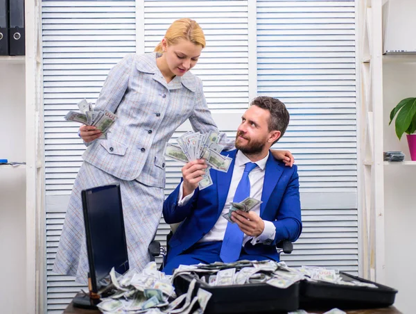 Excited successful businessman opened a box with money and rejoices in profits. Happy young businessman in suit throwing money up while sitting near case full of paper currency.