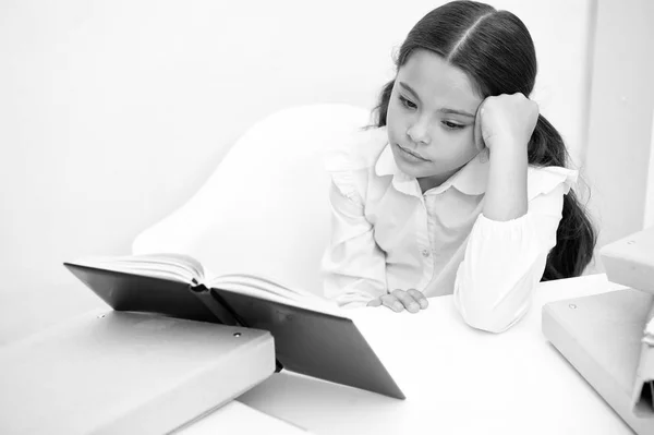 A ler um livro aborrecido. Menina criança lê livro enquanto sentar tabela fundo branco. Estudante estudando e lendo livro. Kid girl escola uniforme entediado rosto indiferente, infelizmente, ler literatura chata — Fotografia de Stock