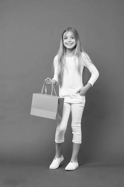 Chica con jersey blanco y pantalones aislados sobre fondo púrpura. Niño con cara alegre sosteniendo bolsa de papel. Niño sorprendido por el regalo, concepto de cumpleaños. Chica con gran sonrisa haciendo compras — Foto de Stock