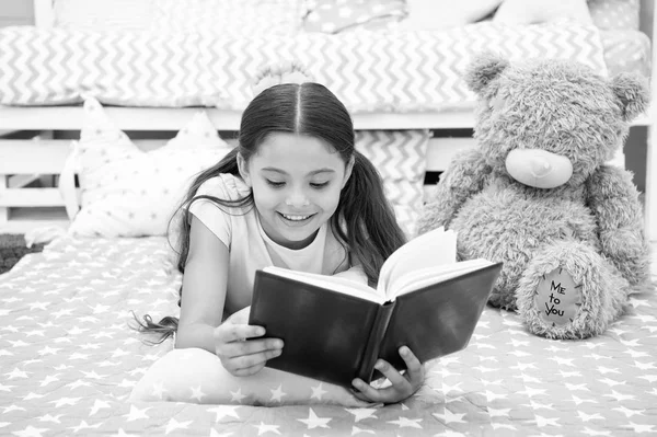 Read fairytale in bed. Girl child lay bed with teddy bear read book. Kid prepare to go to bed. Time for evening fairytale. Girl kid long hair cute pajamas relax and read fairytale book to bear toy