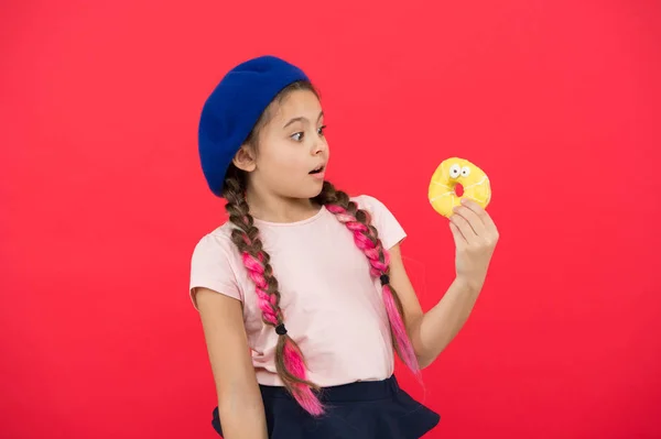 Mädchen mit Baskenmütze halten Donut roten Hintergrund. Kind verspielt Mädchen essen Donut. Gesundheits- und Ernährungskonzept. Süßes Leben. Süßwarengeschäft und Bäckereikonzept. Kid Fan von gebackenen Donuts. leckerer süßer Donut — Stockfoto