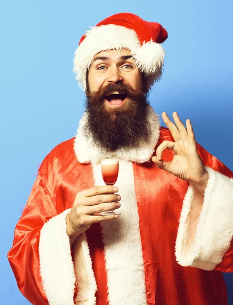 Bonito barbudo santa claus homem com longa barba no sorriso rosto segurando vidro de tiro alcoólico no vermelho natal ou xmas suéter e novo chapéu ano mostrando ok no azul estúdio fundo — Fotografia de Stock