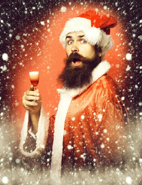 Guapo barbudo hombre de Santa Claus con barba larga en la cara sorprendida sosteniendo vaso de trago alcohólico en navidad o suéter de Navidad y sombrero de año nuevo en fondo rojo estudio — Foto de Stock