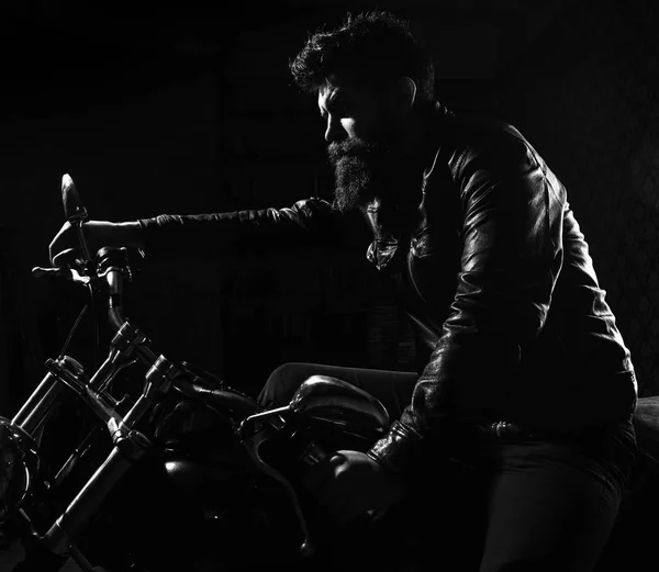 Macho, brutal biker in leather jacket riding motorcycle at night time, copy space. Man with beard, biker in leather jacket sitting on motor bike in darkness, black background. Night racer concept