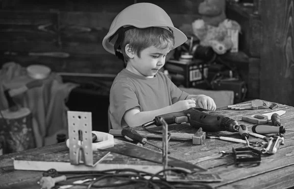 小さな子供のドリルとネジで遊んでボルトします。テーブルに座ってオレンジ ヘルメット側ビュー少年。仕事で少し修理 — ストック写真
