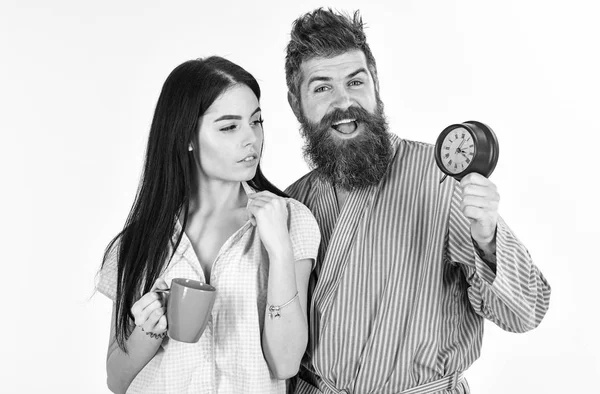 Perfect morning concept. Couple, family woke up on time. Couple in love, young family in pajama, bathrobe stand isolated on white background. Girl with coffee cup, man holds clock in hand — Stock Photo, Image