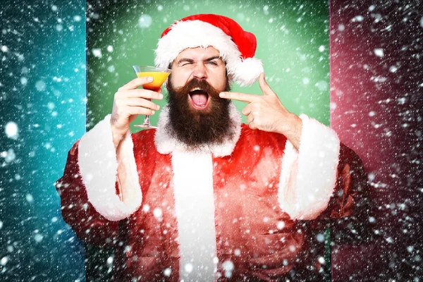 Όμορφος γενειοφόρος Άγιος Βασίλης άνθρωπος με μακριά γενειάδα στο αστείο πρόσωπο κρατώντας ποτήρι αλκοολούχο ποτό τα Χριστούγεννα ή Χριστούγεννα πουλόβερ και το νέο έτος καπέλο δείχνει σε πολύχρωμο φόντο στούντιο — Φωτογραφία Αρχείου
