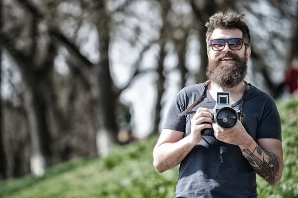 Concepto de fotógrafo. Hombre barbudo hipster fotógrafo mantenga la cámara vintage. Fotógrafo con barba y bigote fotógrafo aficionado fondo de la naturaleza. Hombre con barba larga ocupado con la toma de fotos —  Fotos de Stock