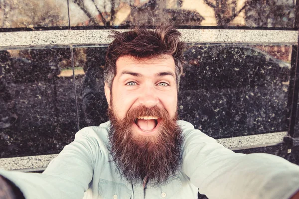 Concepto de vlogging. Hombre, turista con barba y bigote sobre rostro alegre y sonriente, fondo de mármol negro. Hipster, turista con el pelo alborotado y la barba larga mirando a la cámara, tomando foto selfie — Foto de Stock
