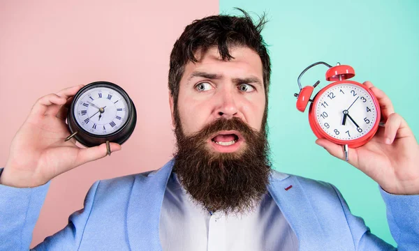Changing time zones affect health. Time zone. Does changing clock mess with your health. Man bearded hipster hold two different clocks. Guy unshaven puzzled face having problems with changing time