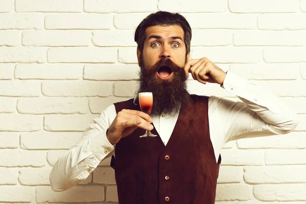 handsome bearded man with long beard and mustache has stylish hair on surprised face holding glass of alcoholic shot in vintage suede leather waistcoat on white brick wall studio background